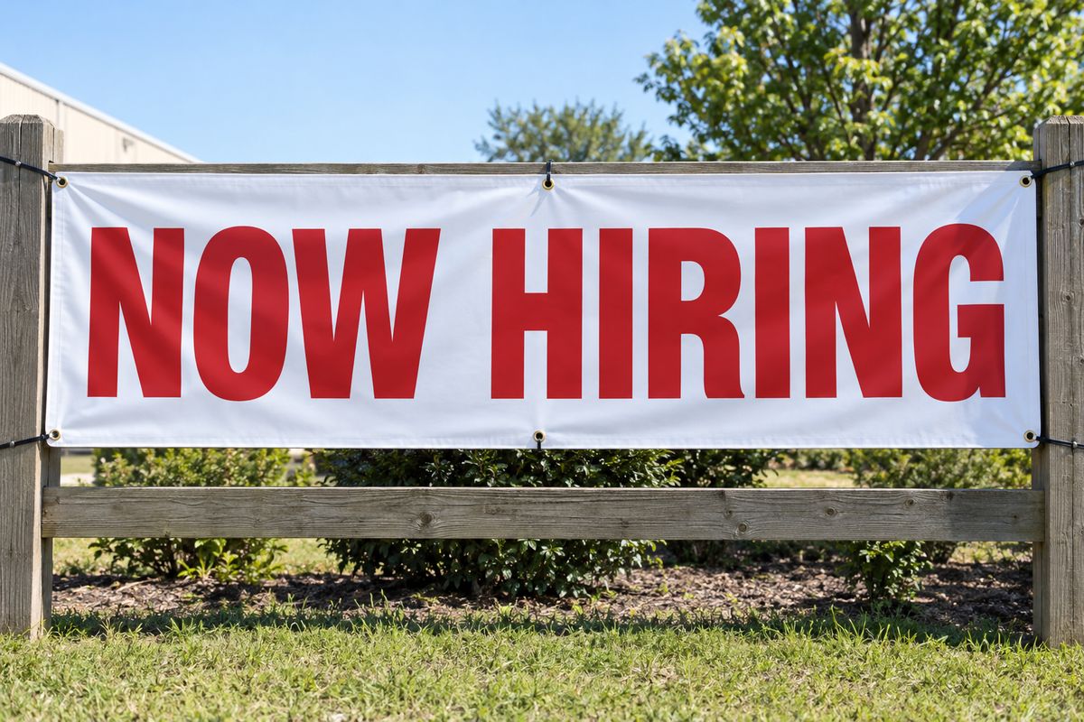 A Now Hiring vinyl banner on a wooden fence.