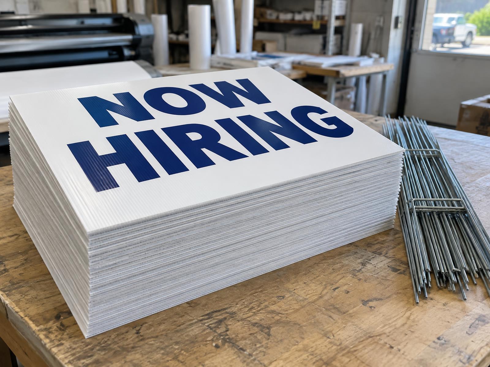 Stack of about 40 freshly printed 18 x 24 in yard signs on a print shop table with a bundle of metal H-stakes beside them, the top sign reading NOW HIRING in navy on white
